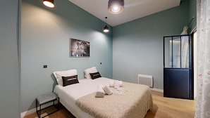 A bedroom with a bed, pillows, a small table, a chair, a radiator, and a window with curtains.