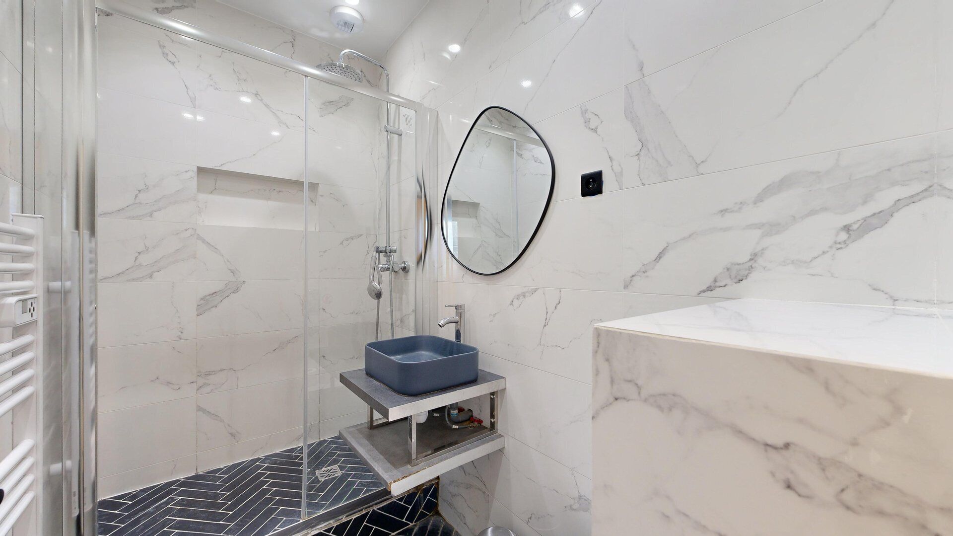 A modern bathroom with a glass-enclosed shower, a marble-tiled wall, a round mirror, and a blue sink.