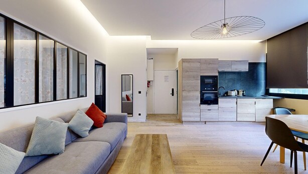 A modern living room with a grey sofa, a wooden coffee table, and a kitchen area with a built-in oven and microwave.