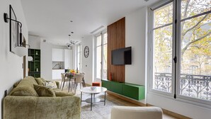A modern living room with a green sofa, a dining area with a table and chairs, and a TV mounted on the wall.