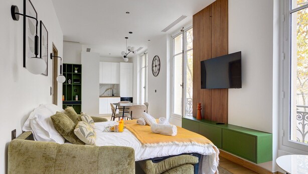 A modern bedroom with a bed, green upholstered furniture, a TV, and a view of the outside.