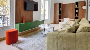 A living room with a green sofa, a round coffee table, and a red ottoman. There is a TV mounted on the wall and a bookshelf with various items.