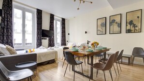 A modern hotel room with a sofa, dining table, and chairs. There is a TV mounted on the wall, a window with curtains, and artwork on the walls. - TBS 47 - Elegant apartment in Le Marais district (Paris)