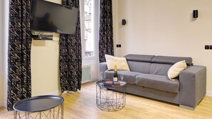 A modern living room with a grey sofa, a flat-screen TV mounted on the wall, a small round table with a bottle of champagne and glasses, and patterned curtains. - TBS 47 - Elegant apartment in Le Marais district (Paris)