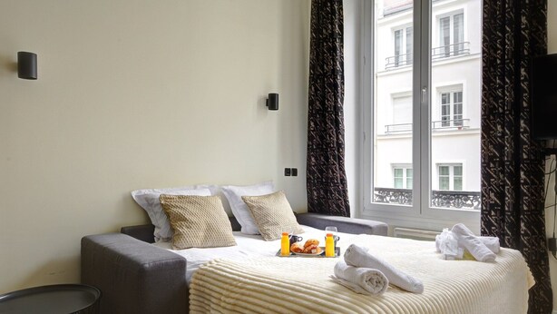 A hotel room with a bed, a grey sofa, a small round table, and a window with curtains. - TBS 47 - Elegant apartment in Le Marais district (Paris)