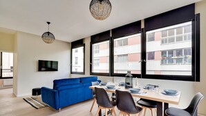 En moderne stue med blå sofa, spisebord med stole og store vinduer med udsigt til bygninger.