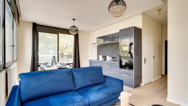 En moderne stue med en blå sofa, et spiseområde med dækket bord og et køkken med lysegrå skabe.