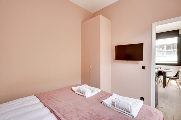 A bedroom with a bed, a TV mounted on the wall, and a door leading to a dining area.