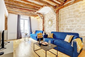 A living room with a blue sofa, a coffee table, a TV, and a stone wall. - TheBlueStay 282 - Cozy Apt w/Terrace - Le Marais (Paris)