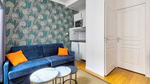 Un espacio habitable compacto con un sofá azul, dos mesitas redondas, una zona de cocina con microondas y una puerta blanca.