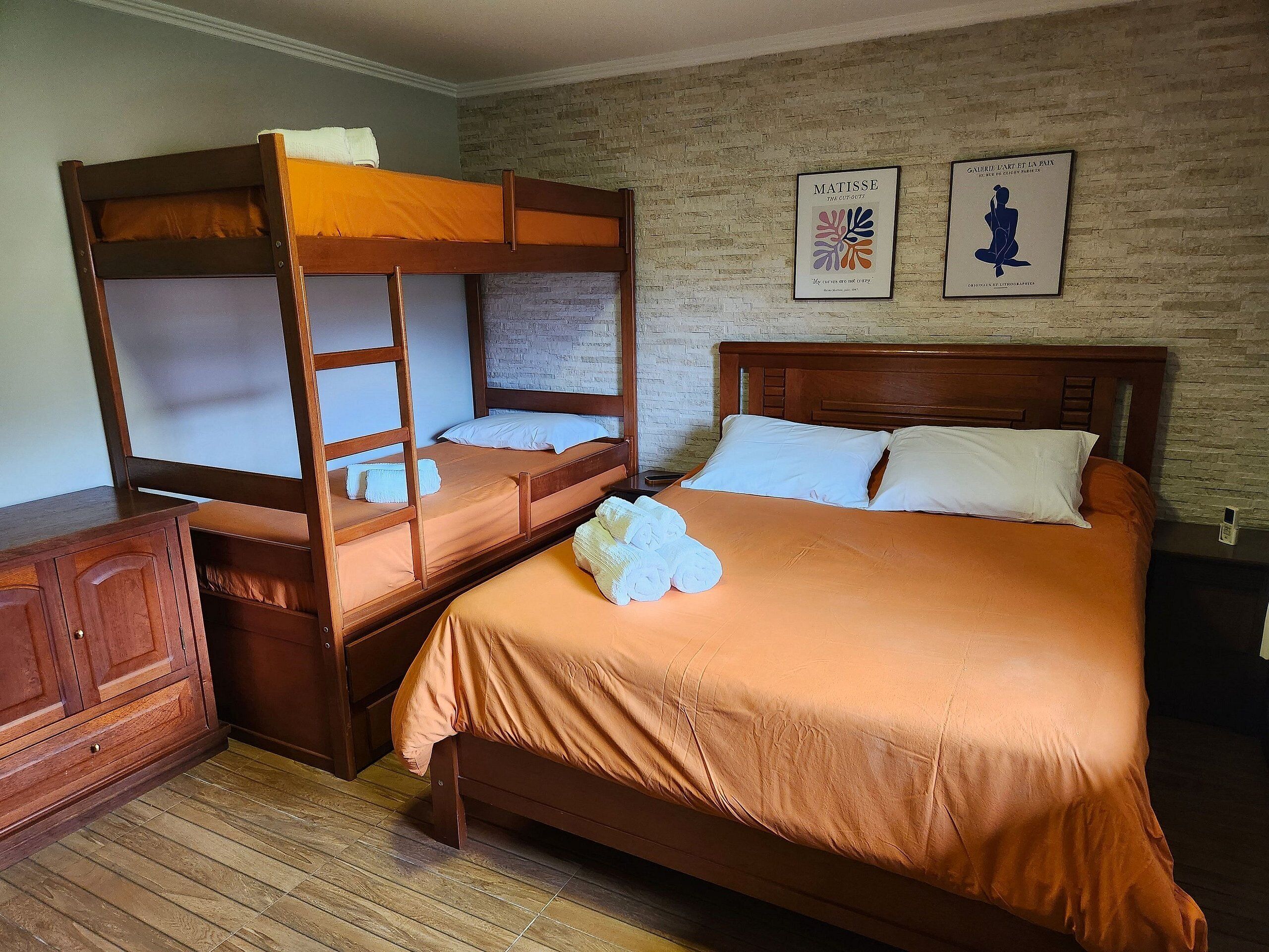 Una habitación con una cama individual, una litera, muebles de madera y dos cuadros en la pared.
