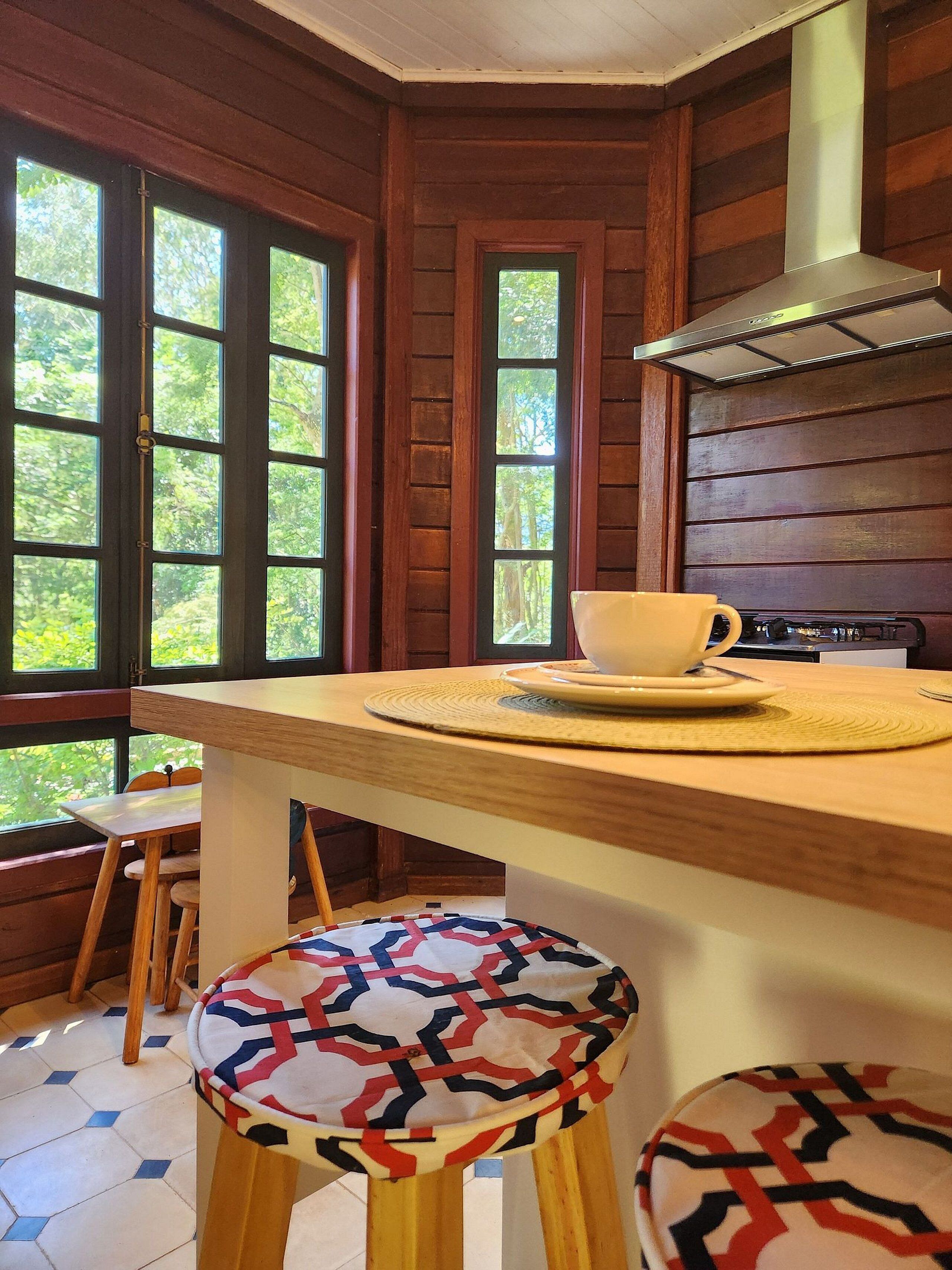 A wooden cabin interior with a kitchen area, a dining table with stools, and large windows allowing natural light.