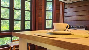 Interior de una cabaña de madera con cocina, una mesa de comedor con taburetes y ventanas amplias que dejan entrar la luz natural.