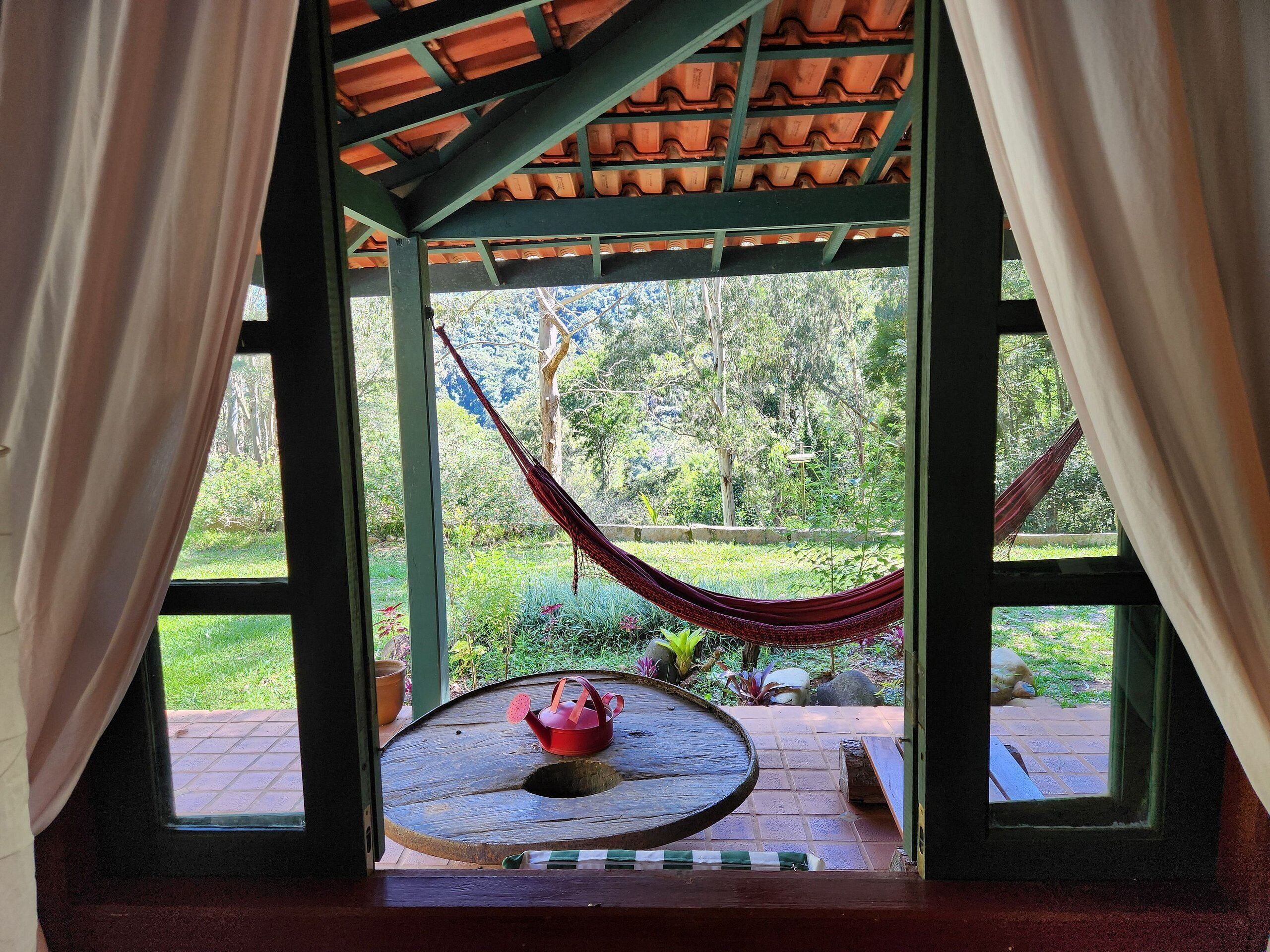 Eine Hängematte, die an einer Holzbalkendecke befestigt ist, ein kleiner Tisch mit einem Teekessel und ein Blick auf einen Garten durch ein blickdichtes Fenster.