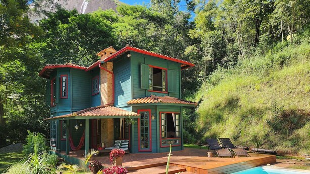 Una casa de dos plantas con piscina, rodeada de exuberante vegetación y una colina.