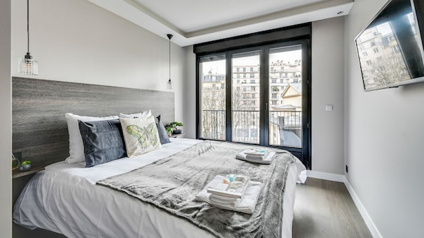 Une chambre moderne avec un grand lit, une vue sur les immeubles par la fenêtre et un téléviseur fixé au mur.