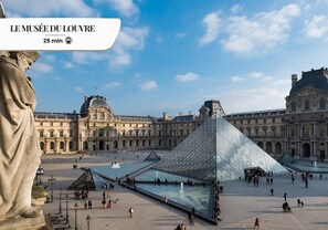 Un'ampia piazza aperta con una struttura piramidale in vetro ben visibile, circondata da edifici in stile classico.