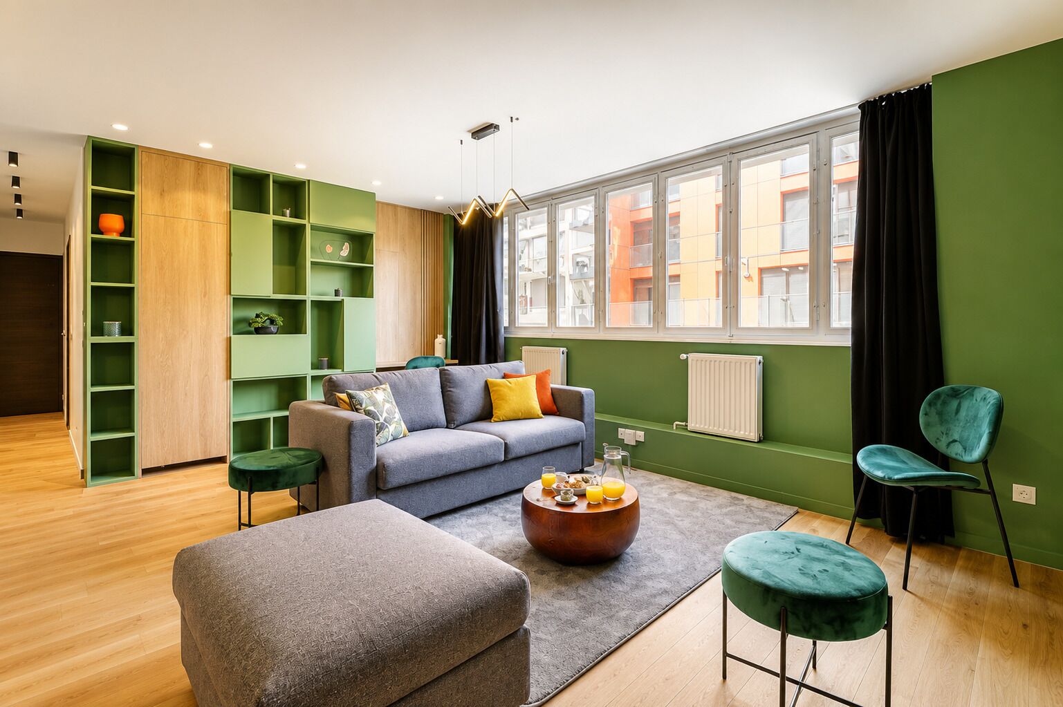 Un salon moderne avec un canapé gris, une table basse et un pouf vert. On y trouve des étagères vertes et une fenêtre avec des rideaux.