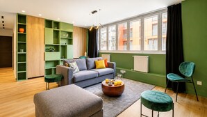 Een moderne woonkamer met een grijze bank, een salontafel en een groene poef. Er staan groene planken en een raam met gordijnen.