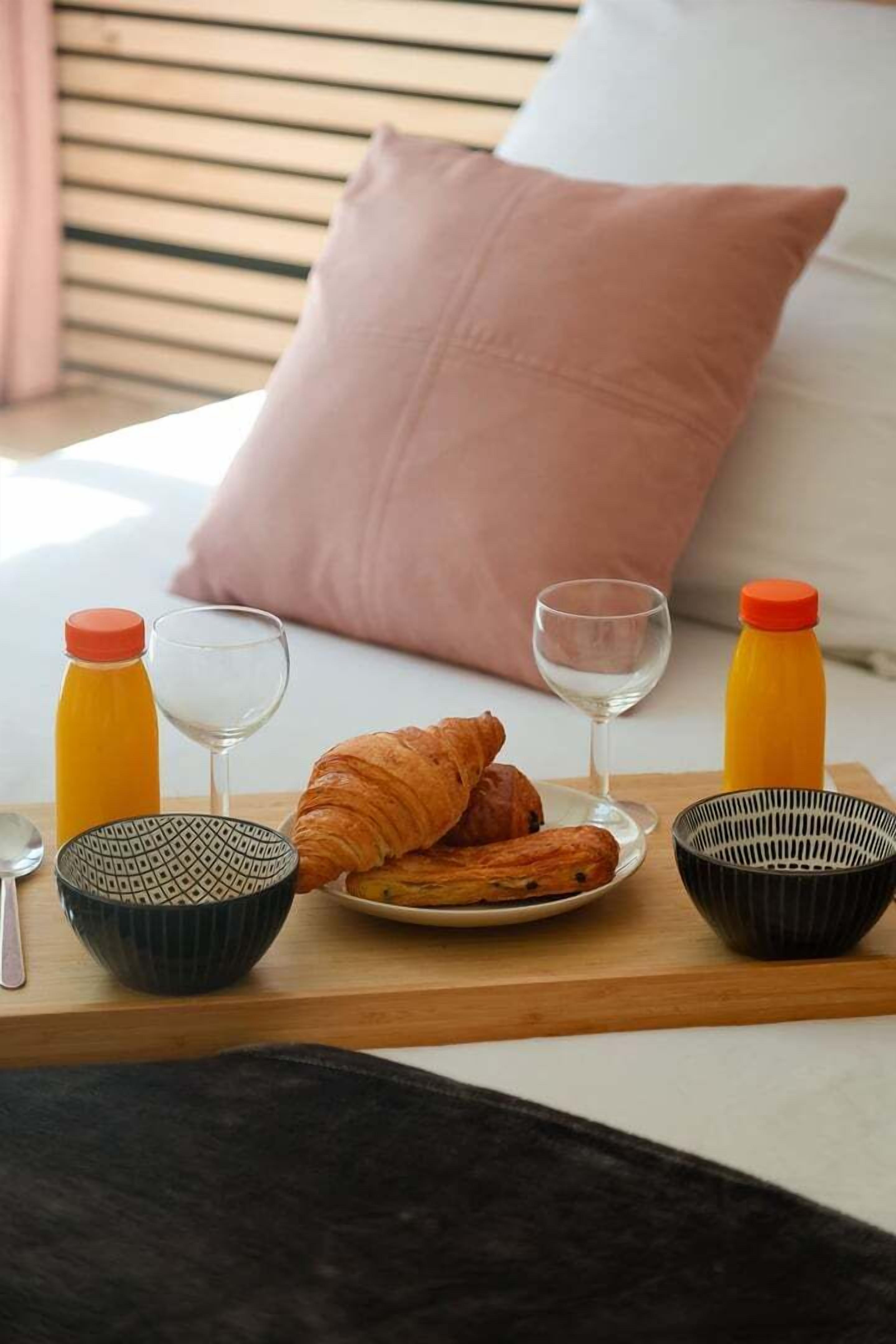 A breakfast tray with croissants, two glasses, a bottle of orange juice, and a bottle of white wine on a bed with a pink pillow.