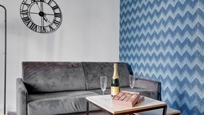 Un sofá gris con un reloj en la pared, una mesa con una botella y vasos, y un papel tapiz estampado.