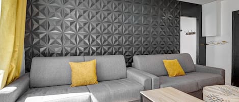 Uma sala de estar moderna com um sofá cinza, duas almofadas amarelas, uma mesa de centro de madeira e um cobertor estampado sobre um banquinho branco.