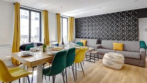 Un moderno comedor con una mesa de madera dispuesta para cuatro personas, rodeada de sillas coloridas. Un sofá gris con cojines amarillos se encuentra frente a una pared con diseño geométrico.