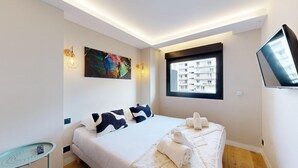 Une chambre moderne avec un lit, une petite table, un tableau, une fenêtre donnant sur des immeubles et un téléviseur fixé au mur.