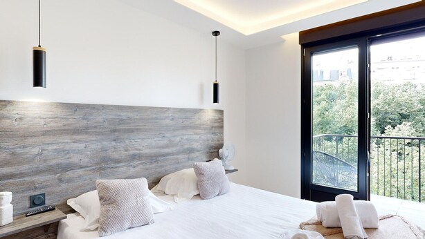 Une chambre moderne avec un grand lit, une tête de lit en bois, un balcon donnant sur les arbres et un luminaire au plafond.