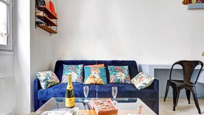 A living room with a blue sofa, floral cushions, a glass coffee table with a bottle and glasses, and a bookshelf with books and decorative items. - Cosy apartment near Opéra Garnier - Suite 222 (Paris)