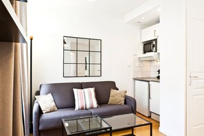 Una sala de estar moderna con un sofá gris, una mesa de centro de vidrio y una zona de cocina con armarios blancos y un microondas.