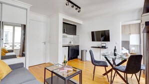 En moderne stue med et sofabord i glas, en grå sofa og et spiseområde med et bord og stole. Et køkken med mørke skabe og et fjernsyn ophængt på væggen er synligt.