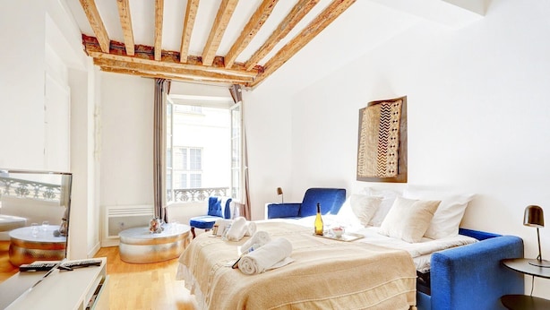 A modern hotel room with a wooden ceiling, a bed with white linens, a blue sofa, a small round table with a bottle and glasses, and a balcony with a view. - TheBlueStay 284 - Charming Apartment - Le Marais (Paris)