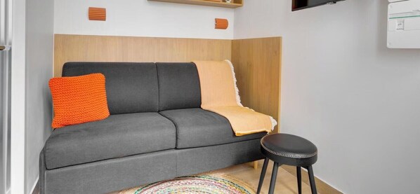 Ein modernes Zimmer mit einem grauen Sofa, einem orangefarbenen Zierkissen, einer beigen Tagesdecke und einem schwarzen Hocker.