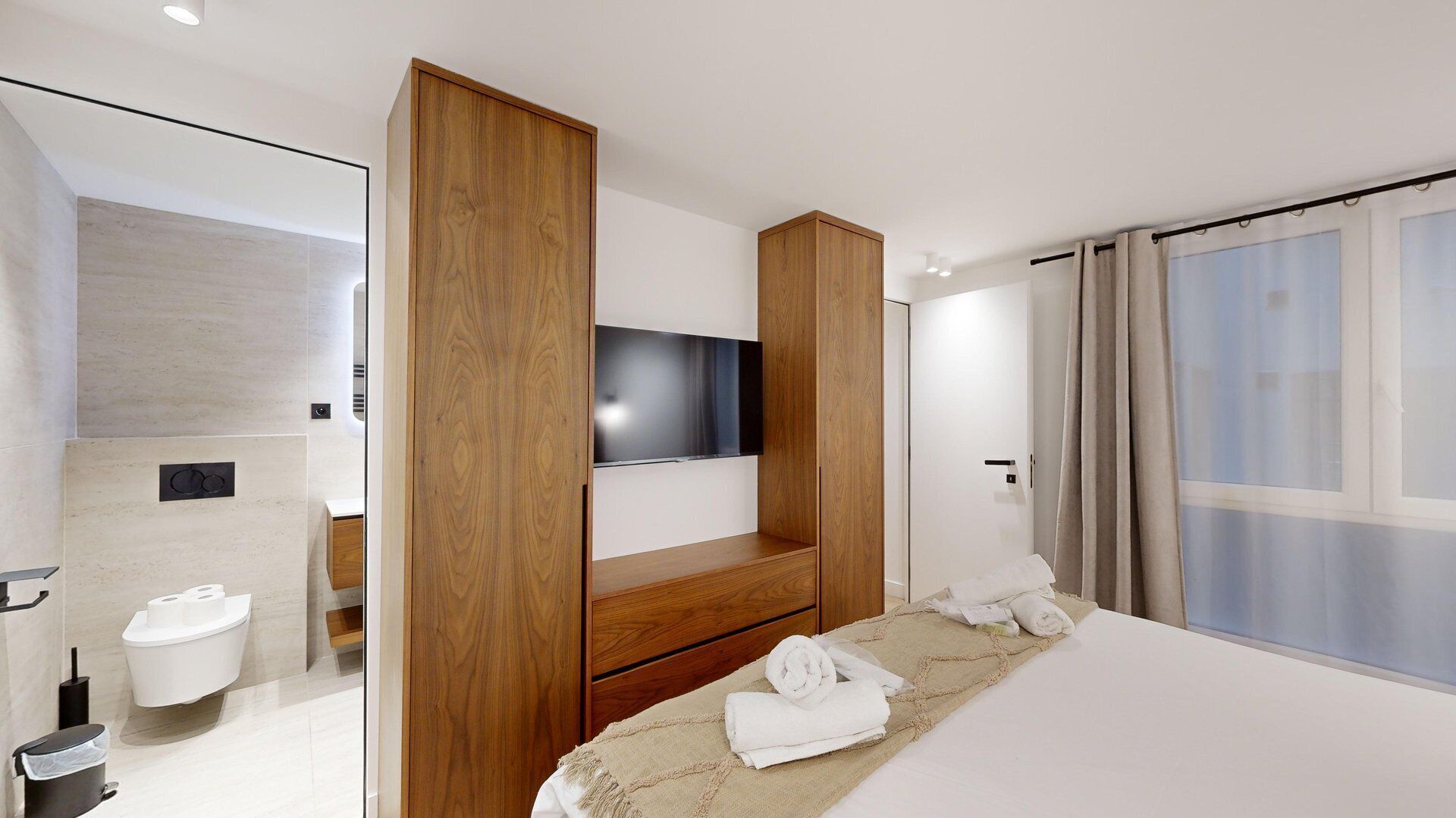 Ein modernes Schlafzimmer mit einem großen Bett, einem an einer Holzvertäfelung montierten Fernseher, einem Badezimmer mit Toilette und Waschbecken sowie einem großen Fenster mit Vorhängen.