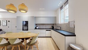 A modern kitchen with white cabinetry, a granite countertop, and a dining area with a wooden table and chairs. - TBS 61 - Pretty suite near Roland-Garros stadium (Paris 16 Passy)
