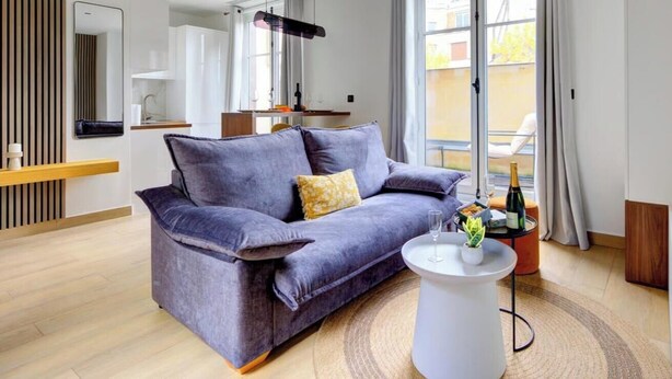 Ein stilvolles Wohnzimmer mit einem grauen Sofa, einem kleinen weißen Beistelltisch mit einer Pflanze und einer Champagnerflasche.