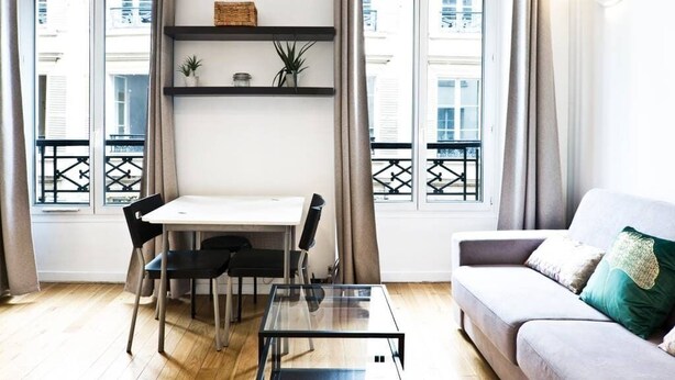 A modern living room with a glass coffee table, a white dining table with chairs, a grey sofa with cushions, and a black bookshelf with decorative items. - TBS 274 - Lovely apartment near Sacré-Coeur (Paris)