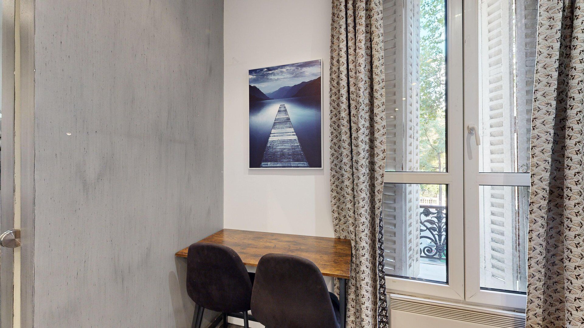 Una habitación con un escritorio de madera, dos sillas, un cuadro enmarco de un muelle y cortinas con un estampado.