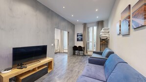 Un salon moderne avec un téléviseur à écran plat posé sur un meuble en bois, un canapé gris, un coin repas avec deux chaises et une fenêtre agrémentée de rideaux à motifs.