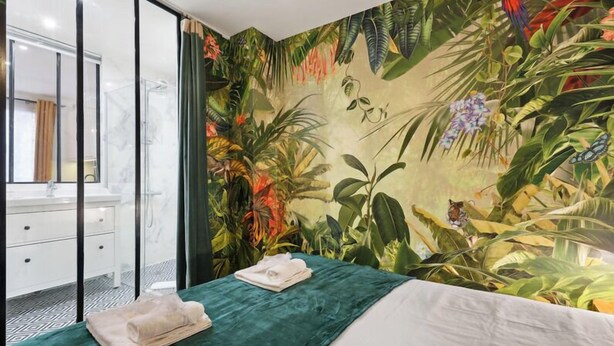 Habitación de hotel con un amplio mural tropical, una cama con cubrecama verde y un baño con cabina de ducha de vidrio.