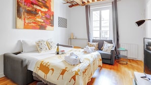 Quarto de hotel com cama, sofá, janela com cortinas, mesa pequena e abajur.