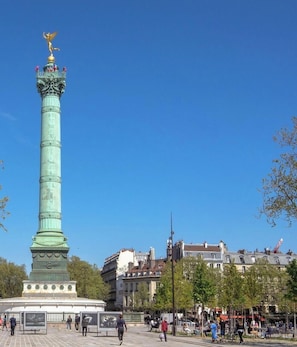Un monumento verde y alto con una estatua dorada en la parte superior, rodeado por una plaza con bancos y árboles.