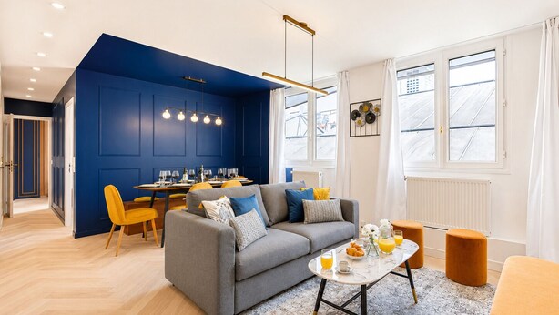 Una sala de estar moderna con un sofá gris, sillas amarillas y un comedor con una mesa dispuesta para dos.