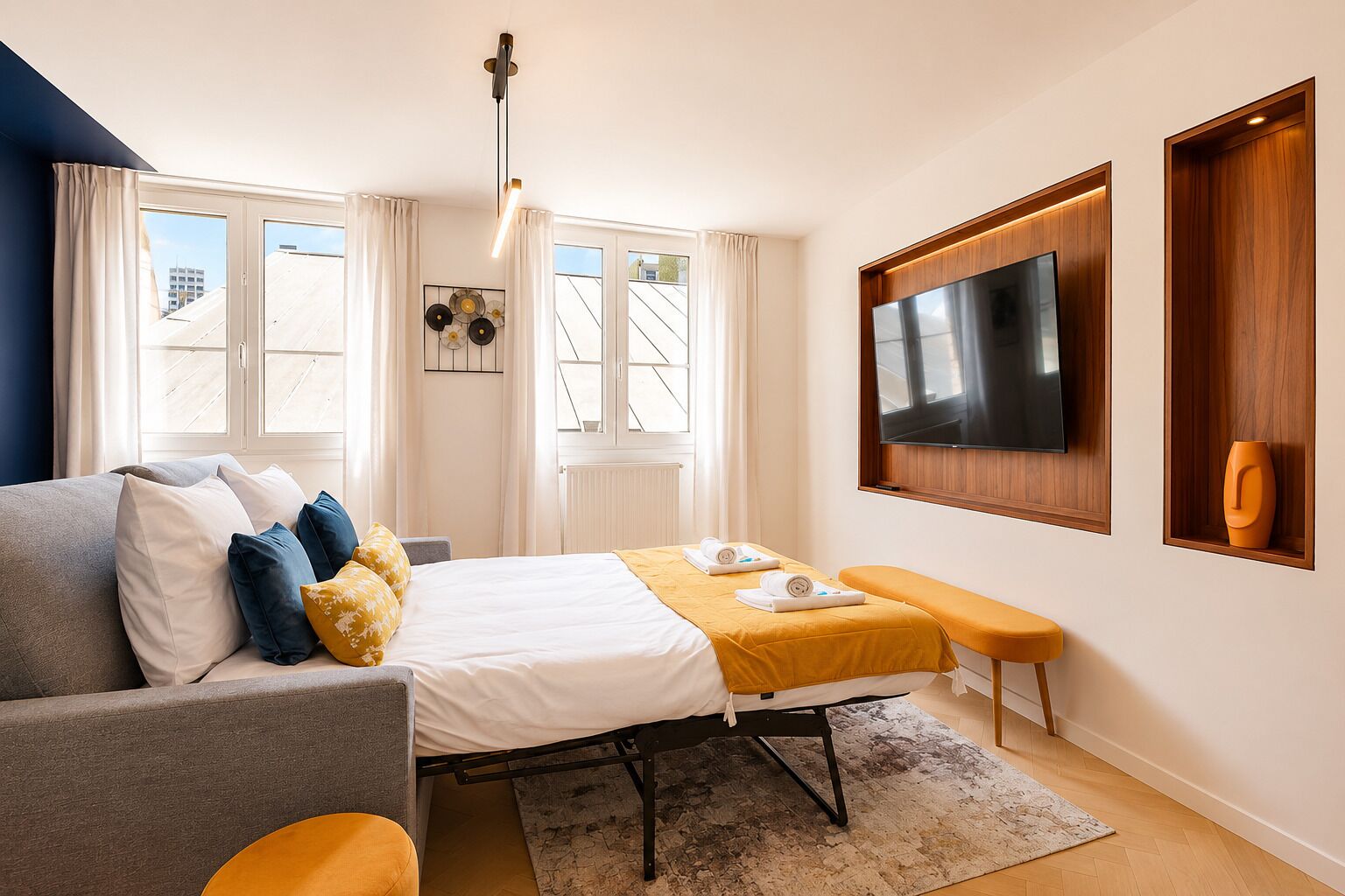 A hotel room with a bed, a sofa, a bench, a TV, and a window with curtains.