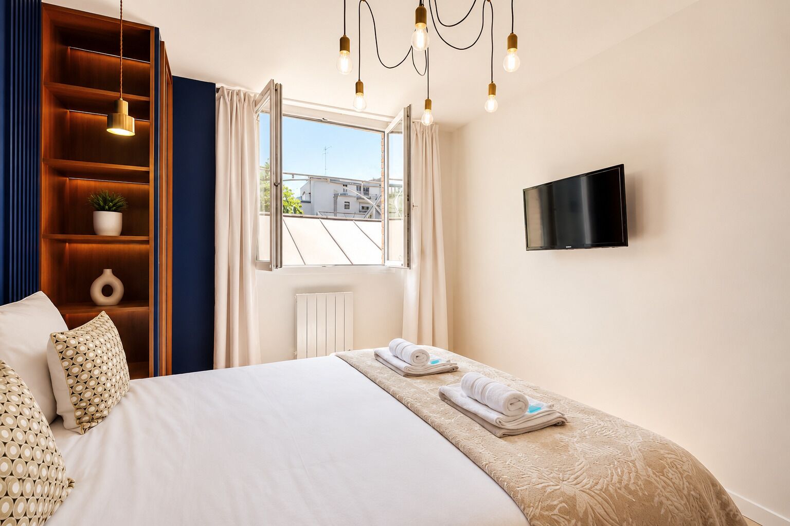 A hotel room with a bed, a TV, a window with curtains, a wooden shelf with decorative items, and a radiator.
