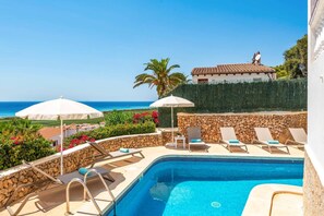A swimming pool with lounge chairs and umbrellas, a stone wall, and a view of the sea.