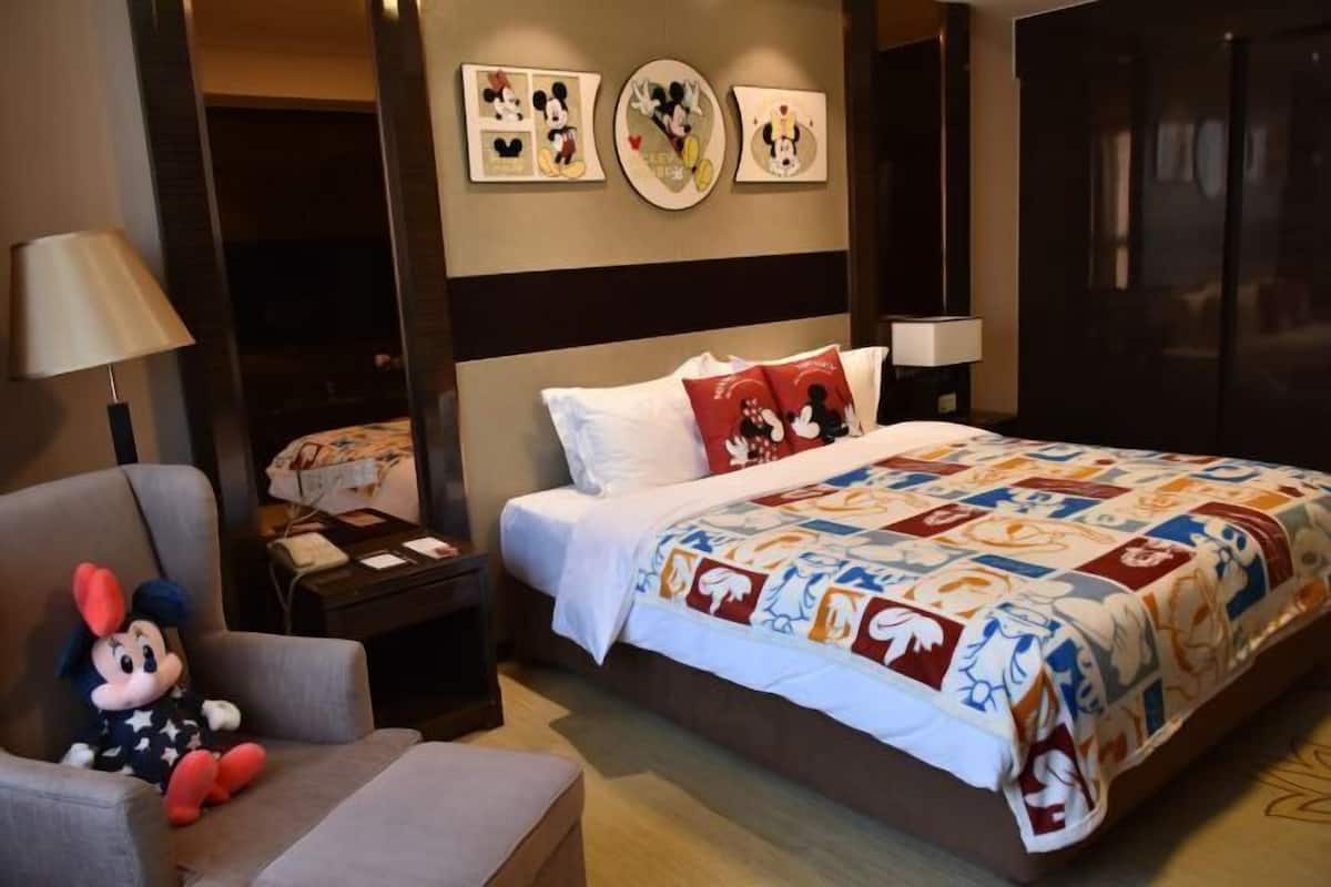 Une chambre d’hôtel avec un lit, un doudou à l’effigie de Mickey Mouse, une lampe et des œuvres d’art mural.