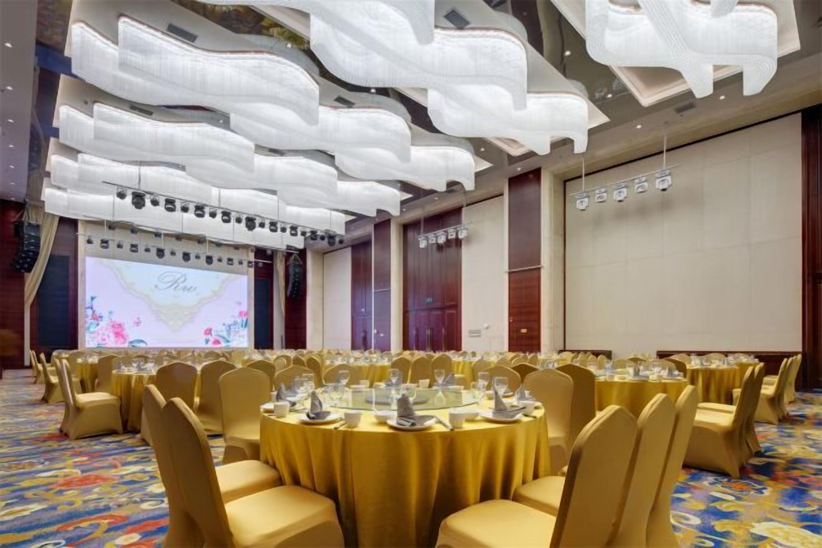 Un salón de banquetes con mesas redondas, sillas y una gran pantalla.
