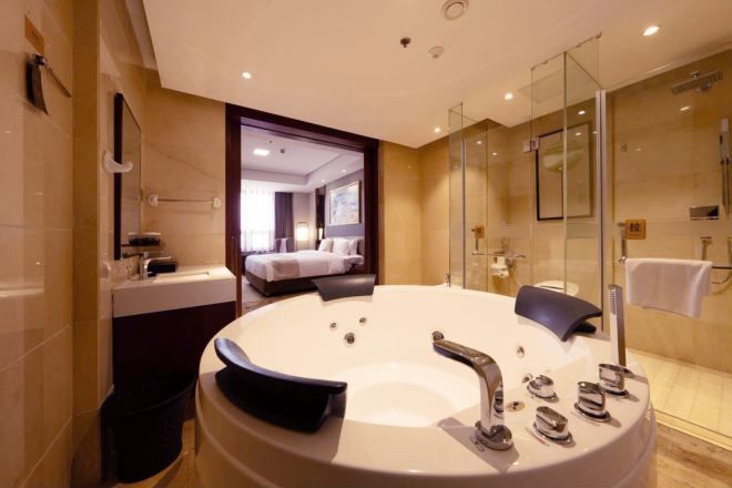 A modern bathroom with a large bathtub, a glass shower enclosure, and a visible bedroom with a bed and a window.